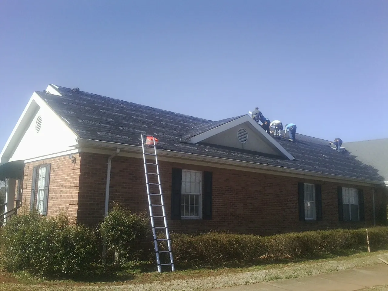 Crew tearing shingles off a brick ranch roof with a ladder set — Emergency Roof Repair work by our crew in Vance
